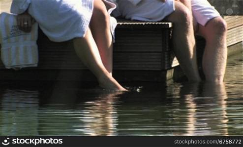 Ein Paar macht Wellness, sitzen im Bademantel auf dem Steg, unterhalten sich und lassen die Beine in Wasser baumeln.