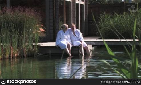 Ein Paar macht Wellness, unterhalten sich, sitzen in BademSntel auf dem Steg und lassen die Beine ins Wasser hSngen.