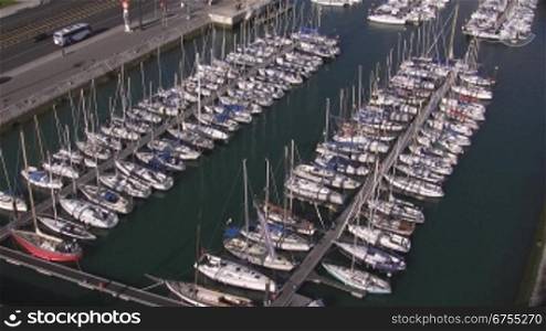 Ein Yachthafen; an Docks angelegte wei?e Segelboote liegen in einem Hafen nebeneinander. Direkt am Hafen eine befahrene Stra?e.
