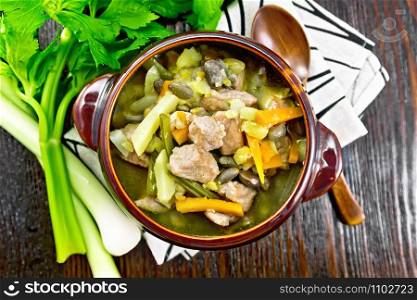 Eintopf soup of pork, celery, beans, carrots and potatoes with leek in a clay bowl on a towel on wooden board background from above