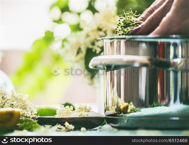 Elder flower syrup or jam cooking. Close up of female hand with Elderflowers and pot on kitchen table at window. Home lifestyle