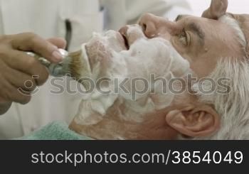 Elderly barber working in old style shop or men&acute;s beauty parlor and shaving client with razor