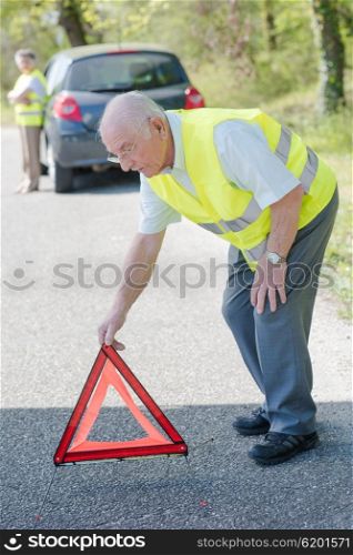 Elderly couple broken down in car, man putting red warning triangle on road