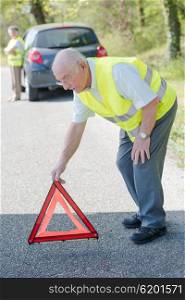 Elderly couple broken down in car, man putting red warning triangle on road