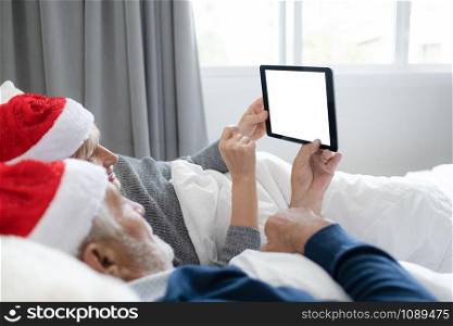 elderly couple caucasian senior man and woman with red hat using internet on tablet with white screen background for christmas festival day in bedroom, retirement love family lifestyle concept