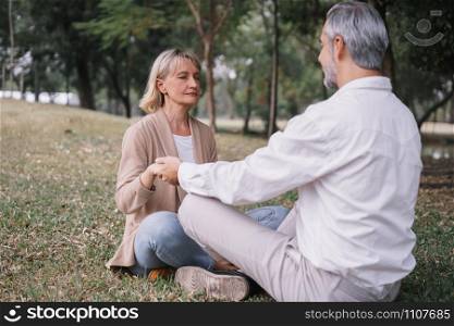 Elderly couple in practicing yoga while sitting holding to hand together in the public park. Concept of relaxation and meditation.