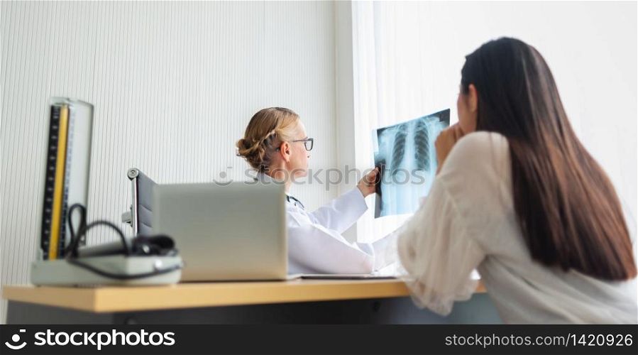 Elderly Female doctor holding and looking to young woman patient X-ray film in medical room at hospital. Diagnosis disease about Lung. Covid-19, Coronavirus.