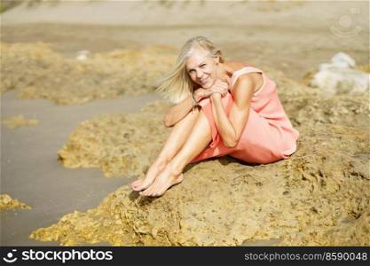 Elderly female sitting on some rocks on the shore of a tropical beach, wearing a nice orange dress. Mature woman enjoying her retirement at a seaside retreat.. Elderly female sitting on some rocks on the shore of a tropical beach, wearing a nice orange dress.