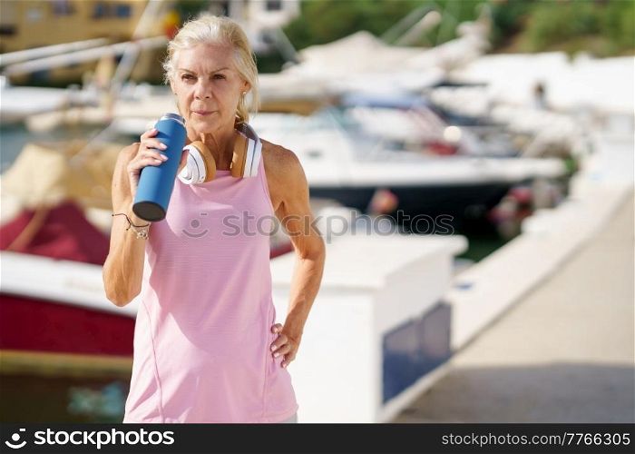 Elderly fit female athlete with headphones on neck drinking water and having break during workout in harbor in summer. Senior sportswoman drinking water during training