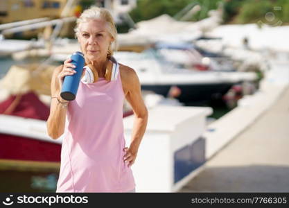 Elderly fit female athlete with headphones on neck drinking water and having break during workout in harbor in summer. Senior sportswoman drinking water during training