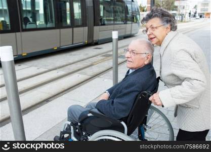 elderly lady pushing husband in wheelchair towards tram