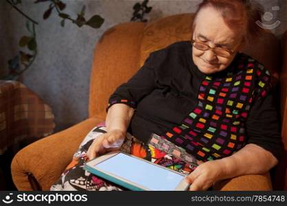 Elderly lady sitting in a comfortable arrnchair in her living room using a tablet computer as she surfs the internet