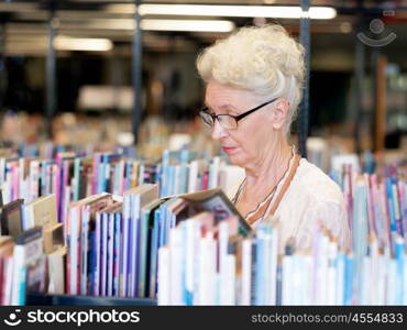 Elderly lady standing next to book shelves in library. Taking her time with new books