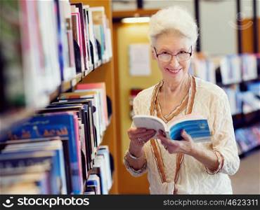 Elderly lady standing next to book shelves in library. Taking her time with new books