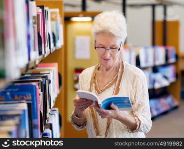 Elderly lady standing next to book shelves in library. Taking her time with new books