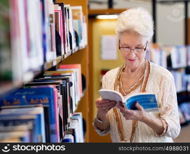 Elderly lady standing next to book shelves in library. Taking her time with new books