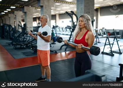Elderly man and female personal trainer, exercise with bar, gym interior on background. Sportive grandpa with woman instructor, workout in sport center. Elderly man and female trainer, exercise with bar