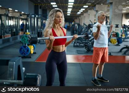 Elderly man and female personal trainer, exercise with bar, gym interior on background. Sportive grandpa with woman instructor, workout in sport center. Elderly man and female trainer, exercise with bar