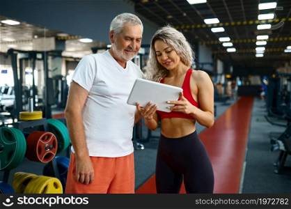 Elderly man and female personal trainer looks on laptop, gym interior on background. Sportive grandpa with woman instructor, training in sport center, old sportsman. Healty lifestyle, health care. Elderly man and female trainer looks on laptop