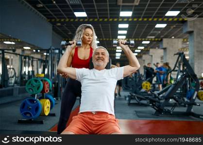 Elderly man doing exercise with dumbbells on bench, female personal trainer, gym interior on background. Sportive grandpa with woman instructor in sport center. Elderly man, exercise with dumbbells on bench