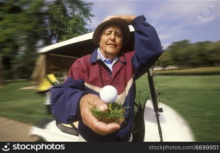 Elderly Man Holding Golf Ball On Tee In Front Of Golf Cart