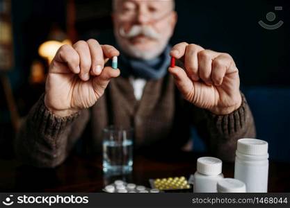 Elderly man holds pills in his hands, home office on background, age-related diseases. Mature senior is ill and being treated in his house. Elderly man holds pills in his hands