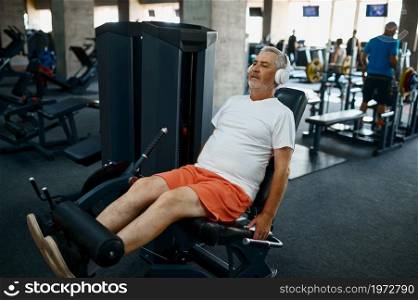 Elderly man in headphones doing abs exercise, gym interior on background. Sportive grandpa on fitness training in sport center. Healty lifestyle, health care, old sportsman. Elderly man in headphones doing abs exercise