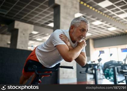 Elderly man in sportswear doing abs exercise, gym interior on background. Sportive grandpa on fitness training in sport center. Healty lifestyle, health care, old sportsman. Elderly man in sportswear doing abs exercise, gym
