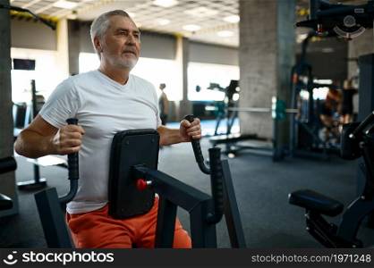 Elderly man in sportswear on exercise machine, gym interior on background. Sportive grandpa on fitness training in sport center. Healty lifestyle, health care. Elderly man in sportswear on exercise machine, gym