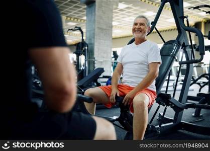Elderly man poses on exercise machine, gym interior on background. Sportive grandpa on fitness training in sport center. Healty lifestyle, health care, old sportsman. Elderly man poses on exercise machine in gym