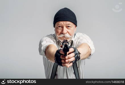 Elderly man poses with gun on grey background, gangster. Mature senior in hat holds weapon, robber in old age. Elderly man poses with gun on grey background