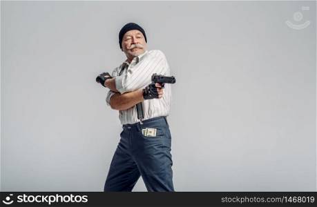 Elderly man poses with gun on grey background, gangster. Mature senior in hat holds weapon, robber in old age. Elderly man poses with gun on grey background