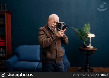 Elderly man poses with old film camera in home office. Bearded mature senior in living room, old age businessman. Elderly man poses with old film camera