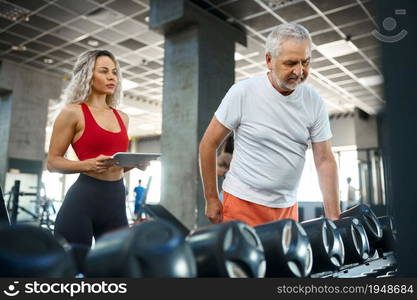 Elderly man takes dumbbells, female personal trainer holds laptop, gym interior on background. Sportive grandpa with woman instructor, training in sport center, old sportsman. Elderly man takes dumbbells, female trainer, gym