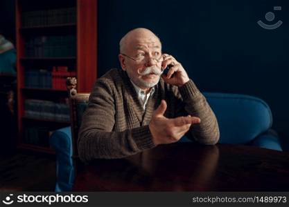 Elderly man talking by mobile phone in home office. Bearded mature senior in living room, old age businessman. Elderly man talking by mobile phone in home office