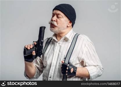 Elderly man with gun isolated on grey background, gangster. Mature senior in hat holds weapon, robber in old age. Elderly man with gun isolated on grey background