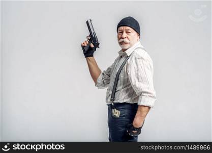 Elderly man with gun isolated on grey background, gangster. Mature senior in hat holds weapon, robber in old age. Elderly man with gun isolated on grey background