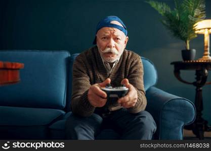 Elderly man with joystick plays a game console on couch. Bearded mature senior poses in living room, old age people leisures. Elderly man with joystick plays a game console