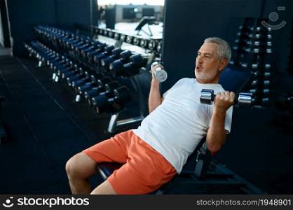 Elderly man, workout with dumbbells on bench, gym interior on background. Sportive grandpa on fitness training in sport center. Healty lifestyle, health care, old sportsman. Elderly man, workout with dumbbells on bench, gym