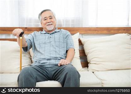 elderly old senior man sitting, resting his hands on wooden walking stick sitting on couch in living room in house after retirement.