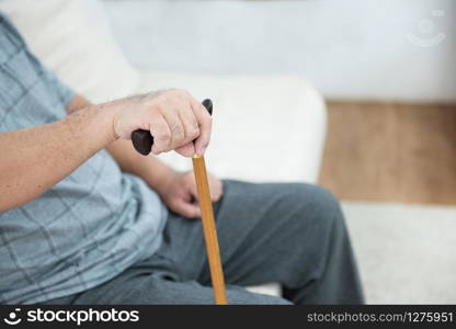 elderly old senior man sitting, resting his hands on wooden walking stick sitting on couch in living room in house after retirement.