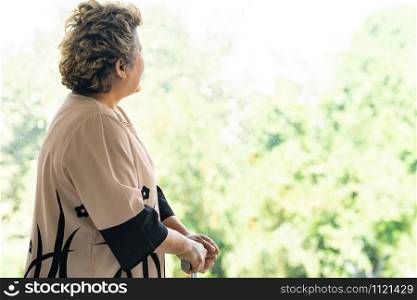Elderly senior female using walking stick or walking cane staff standing beside window in domestic living room - recovery and rehabilitation concept