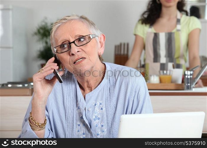 Elderly woman chatting on the phone whilst young helper works in the kitchen