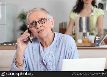 Elderly woman chatting on the phone whilst young helper works in the kitchen
