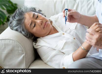 Elderly woman having cold during lie down on couch while Senior man checking temperature of his wife with digital thermometer in the house. Old female having fever. Illness, Disease, Coronavirus, virus, Covid, Spread, Quarantine, Carer.
