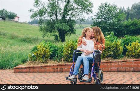 Elderly woman in a wheelchair kissing her daughter in the garden. Woman in a wheelchair kissing her daughter