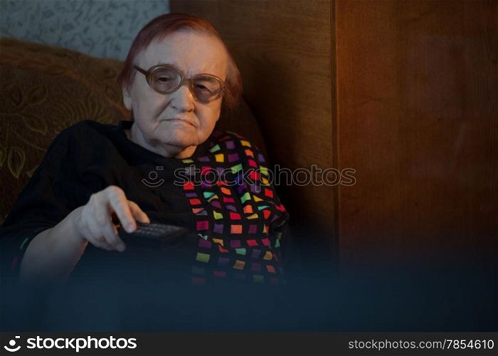 Elderly woman in glasses sitting on the sofa and changing channels on TV with remote control