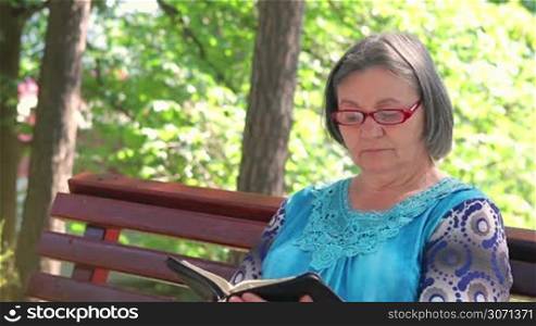 Elderly woman reading bible outdoors on the bench.
