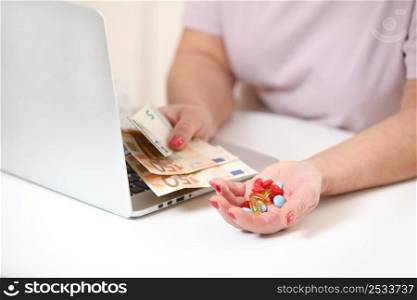 Elderly woman with laptop holding money, euro cash and pills on white table, copy space. Concept of treatment cost, medicine price, health insurance budget.. Elderly woman with laptop holding money, euro cash and pills on white table, copy space. Concept of treatment cost, medicine price, health insurance budget