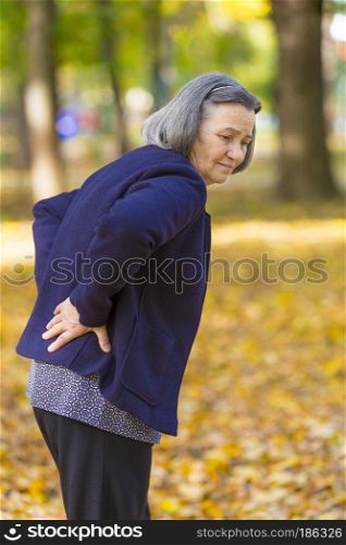 Elderly woman with lower back pain in autumn city park. Unhappy senior woman while feels pain from backache walking in autumn park.